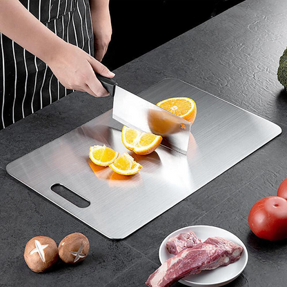 Person cutting oranges on a metal cutting board with vegetables and meat on a dark surface
